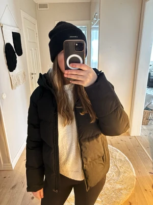 Svart pufferjacka med dragkedja - Säljer en svart pufferjacka med huva och dragkedja framtill. Jackan har en clean look, är vadderad och har långa ärmar. Perfekt för kalla dagar när du vill vara både varm och stilren. Köpt för 800kr på Stadium förra året, endast använd ett fåtal gånger. 