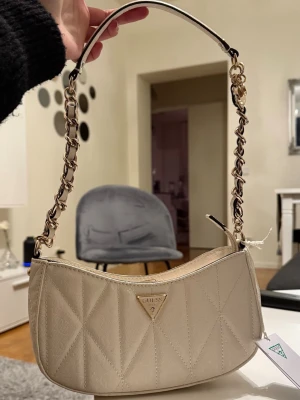 Off white väska från Guess - Säljer en snygg off white axelväska från Guess med quiltad design och guldfärgad kedja på axelbandet. Väskan har Guess-logga i metall framtill och dragkedja upptill. Perfekt storlek för det viktigaste och ger en lyxig vibe till din outfit.