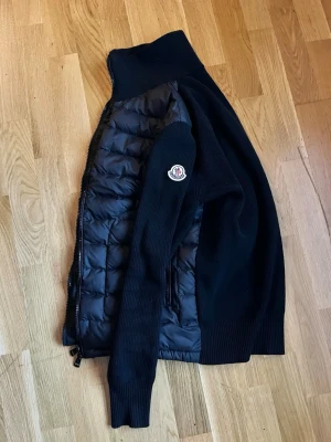 Svart Moncler cardigan jacka S - Säljer en svart cardigan jacka från Moncler i storlek S funkar även XS. Jackan har quiltad front i polyester och stickade ärmar samt rygg. Dragkedja framtill och diskret Moncler-logga på ena ärmen. Perfekt för dig som vill ha en clean och stilren look.