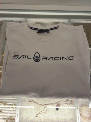 Vit sweatshirt från Sail Racing - Säljer en clean vit sweatshirt från Sail Racing med svart logga tryckt över bröstet. Klassisk rund halsringning med ribbade muddar vid ärmslut och nederkant. Perfekt för dig som gillar stilren design och sportiga vibes.