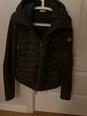 Moncler Cardigan - Säljer en mörkgrön Cardigan från Moncler med stickade ärmar och huva. Jackan har dragkedja framtill, ribbade muddar och Moncler-logga på ärmen. Perfekt för kyliga dagar och riktigt snygg till höst- och vinterlooken.