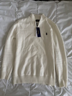 Vit half zip tröja från Polo Ralph Lauren - Vit stickad tröja från Polo Ralph Lauren med half zip och ribbade muddar. Klassisk logga broderad på bröstet och hög krage. Perfekt för lager på lager under höst och vinter. Snygg och enkel att matcha till det mesta.