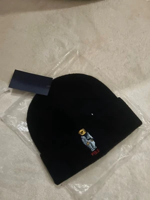 Svart mössa från Polo Ralph Lauren - Svart mössa från Polo Ralph Lauren med broderad björn på framsidan. Klassisk design med uppvikt kant och diskret logga i rött under björnen. Perfekt accessoar för kyliga dagar.