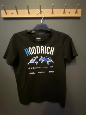 Svart HOODRICH t-shirt med tryck XL - Svart t-shirt från HOODRICH i storlek XL för åldrar 13-15 år. Framsidan har ett stort tryck med texten HOODRICH i vitt och blått samt en världskarta i blå toner. Klassisk rund hals och korta ärmar. Perfekt för dig som gillar streetwear och snygga prints.