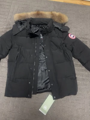 Svart dunjacka från Canada Goose - Säljer en svart dunjacka från Canada Goose med pälskantad huva och klassisk patch på ärmen. Jackan har dragkedja och knappar framtill, stora fickor och är perfekt för kalla vinterdagar. Insidan är fodrad i svart och materialet är slitstarkt och varmt.