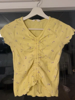 Gul blommig topp från H&M  - Supersöt gul topp från H&M med små lila blommor och ribbad struktur. Toppen har v-ringning, korta ärmar och snygg snörning framtill som ger en cool rynkad effekt. Perfekt för vår och sommar när du vill ha något färgglatt och bekvämt.