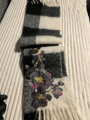 Rutig ullväst med broderade blommor - Säljer en riktigt snygg väst i tjock ullmix med stora svarta och vita rutor. Västen har broderade blommor i grått, brunt, rosa och gult som ger en unik touch. Långa fransar nertill för extra stilpoäng. Perfekt för dig som vill sticka ut lite extra.