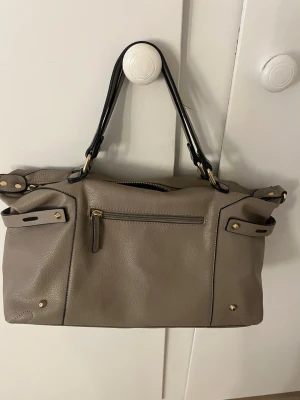 Beige axelväska med gulddetaljer - Säljer en beige axelväska i skinnimitation med svarta handtag och snygga guldfärgade detaljer. Väskan har dragkedja upptill, ytterfack med dragkedja och ett mönstrat innerfoder. Perfekt storlek för allt du behöver ha med dig.