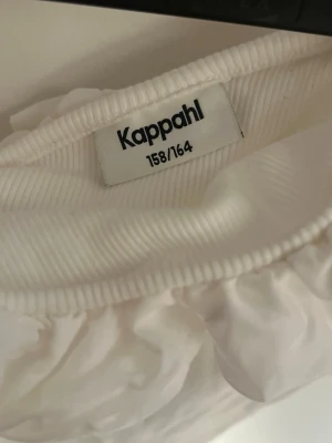 Vit oneshoulder topp med volang Kappahl - Säljer en vit oneshoulder topp från Kappahl i ribbad bomull. Toppen har en bred volang längs axelbandet som ger en söt och trendig look. Perfekt för sommarens alla tillfällen och lätt att matcha med jeans eller kjol.