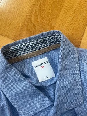 Blå skjorta från Devred 1902 XL - Snygg blå skjorta från Devred 1902 i storlek XL. Skjortan har långa ärmar med gråa armbågslappar, knappar framtill och ett diskret mönstrat tyg på insidan av kragen. Perfekt för dig som vill ha en chill men ändå stilren look.
