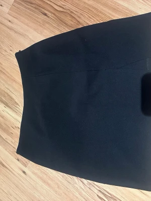 Svart kortkjol från NA-KD - Säljer en svart, stilren kortkjol från NA-KD. Kjolen har en rak passform, slits på sidan och diskret dragkedja i sidan för enkel påklädning. Materialet känns mjukt och följsamt, perfekt till många olika looks.