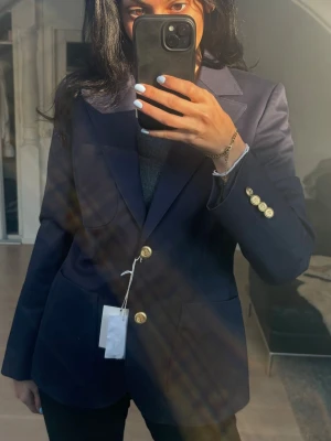 Mörkblå kavaj med guldknappar BOOMERANG - Snygg mörkblå kavaj med klassisk passform och markerade axlar. Kavajen har stora framfickor och stilrena guldknappar både framtill och vid ärmsluten. Perfekt för dig som vill ha en tidlös och dressad look. Materialet känns slätt och håller formen snyggt.