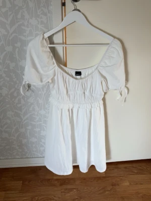 Vit kortklänning från Gina Tricot - Supersöt vit kortklänning från Gina Tricot med puffärmar och knytband vid ärmsluten. Klänningen har en smockad midja och lätt rynkade detaljer,Perfekt för sommarens alla tillfällen!Säljs pågrund av att den inte kommer till användning❤️