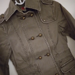 Militärgrön vårjacka / sommarjacka Strlk: XS. Fint skick. - Militärgrön vårjacka / sommarjacka. Stängs med knappar och har 2 fickor. Strlk: XS.   Material: Kraftigare bomullstyg.  Sparsamt använd, fint skick, felfri.  FRAKT TILLKOMMER. 