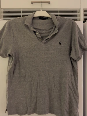 Grå pikétröja Polo Ralph Lauren L - Klassisk grå pikétröja från Polo Ralph Lauren i storlek L, slim fit. Tröjan har korta ärmar, krage med tre knappar och den ikoniska broderade loggan på bröstet. Tillverkad i mjuk bomull för en bekväm känsla och stilren look.