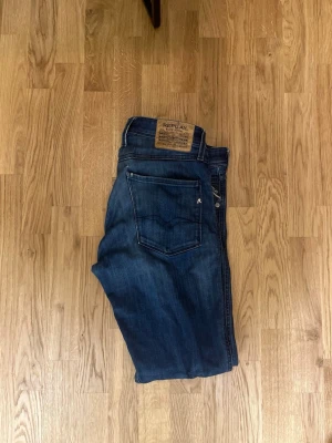 Replay Anbass blå jeans, storlek 32 - Säljer ett par snygga Replay Anbass jeans i klassisk blå tvätt. Jeansen har fem fickor, normal passform och raka ben. Märkespatch bak i midjan och coola slitningar framtill. Perfekta till sneakers eller boots för en avslappnad look.