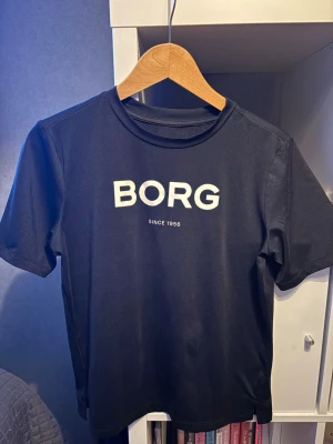 Svart Björn Borg träningst-shirt S - Svart t-shirt från Björn Borg med stor vit logga 'BORG' och texten 'Since 1956' på bröstet. Klassisk rund hals och korta ärmar. Tillverkad i mjuk polyester som känns skön mot huden. Perfekt för dig som gillar sportig och clean stil. Använt få gånger 