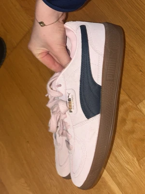 Puma Palermo sneakers i rosa mocka - Säljer ett par Puma Palermo sneakers i ljusrosa mocka med mörkblå sidodetalj och brun sula. Skorna har klassisk snörning och rund tå. Perfekta för dig som vill ha en clean och trendig look med retrovibe.