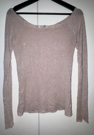 Glittrig beige långärmad topp Design by Si - Säljer en beige långärmad topp från Design by Si med glittrigt lurexmaterial. Toppen har en bred, något offshoulder ringning och är figurnära i passformen. Materialet är mjukt och stretchigt med en skimrande finish som ger en extra touch.