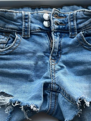 Blå jeansshorts med rå kant - Säljer ett par ljusblå jeansshorts från Gina med låg midja, dubbla knappar och dragkedja. Shortsen har slitna detaljer, råa kanter och klassiska fickor både fram och bak. Perfekta för sommardagar och ger en avslappnad vibe.