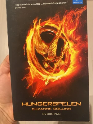 Hungerspelen av Suzanne Collins - Hungerspelen är en spännande dystopisk roman av Suzanne Collins. Omslaget är svart med en eldfängd guldfågel och ring, och texten är i guld. Detta är en MånPocket-utgåva och boken är på svenska. Perfekt för dig som gillar action och äventyr.