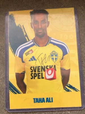 Samlarbild Taha Ali Sverige - Unik samlarbild med Taha Ali i svenska landslagets gula matchtröja. Kortet har en äkta  autograf och tydliga blå och gula färger, samt texten 'SVENSKA SPEL' på framsidan. Perfekt för dig som gillar fotboll och vill ha något speciellt i din samling. 