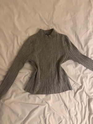 Grå ribbad croppad polotopp - Säljer en ljusgrå ribbad polotopp med lång ärm och croppad passform. Toppen är figurnära och har en hög krage som ger en clean look. Perfekt att styla med både jeans och kjol för en trendig outfit.
