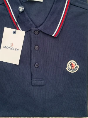 Marinblå pikétröja från Moncler - Snygg marinblå pikétröja från Moncler med klassisk krage och tre knappar framtill. Kragen har röda och vita ränder som detalj och bröstet pryds av Monclers ikoniska logotyp. Perfekt för en stilren och sportig look.