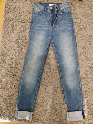 Blå jeans från Lindex med uppvik - Snygga blå jeans från Lindex med klassisk femficksmodell och guldfärgade knappar. Jeansen har uppvikta benslut och en lätt tvättad look med slitningar på låren. Perfekta för en avslappnad och trendig stil.