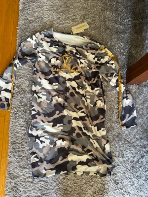 Camouflageklänning med guldtryck - Säljer en långärmad klänning med camouflage-mönster i grå, svart och beige toner. Klänningen har ett guldfärgat tryck och text på bröstet samt dekorativa guldfärgade band längs ärmarna. Materialet känns mjukt och stretchigt, perfekt för en cool och avslappnad stil. Skriv privat för fler bilder eller om du har några frågor💗