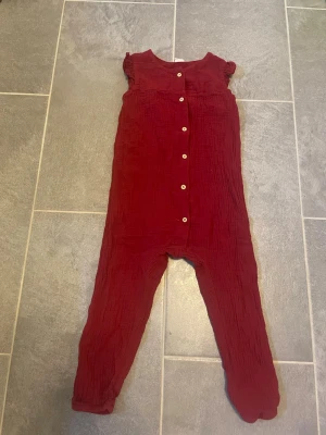 Vinröd jumpsuit med knappar - Vinröd jumpsuit i mjukt, krinklat tyg med långa ben och korta volangärmar. Knäppning framtill med vita knappar. Perfekt för dig som vill ha en bekväm och stilren look. Materialet känns luftigt och skönt mot huden.