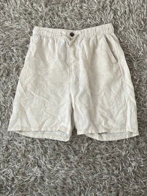 Beiga linneshorts med resårmidja - Snygga och luftiga beiga shorts i linne med elastisk midja och knapp framtill. Shortsen har två sidofickor och en avslappnad passform, perfekta för varma dagar.