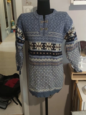 Blå mönstrad stickad tröja - Mysig blå stickad tröja med traditionellt nordiskt mönster i vitt, beige och mörkblått. Tröjan har långa ärmar, ribbade muddar och dekorativa spännen vid halsen. Perfekt för kalla dagar och ger en klassisk vinterlook.