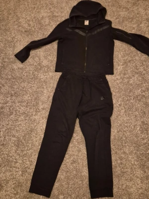 Svart nike tech set - Svart set från Nike skön byxor med dragsko i midjan och muddar vid bensluten. En tröja med snygga detaljer, har en klassisk sportig look och är tillverkade i mjukt material. Perfekta för chill eller träning.