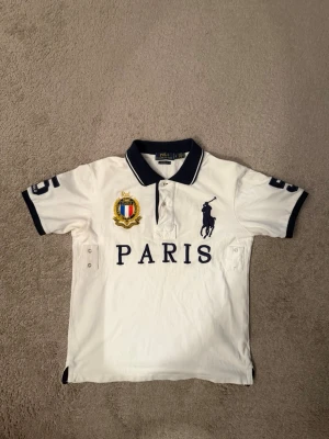 Polo Ralph Lauren Big Pony Paris Polo White - Vit pikétröja från Polo Ralph Lauren med mörkblå detaljer, stor logga och texten PARIS framtill. Nummer 5 på ryggen och ärmarna, samt broderad patch på bröstet. Klassisk krage och korta ärmar. Perfekt för en sportig och stilren look.