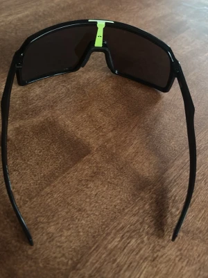 Svarta sportiga solglasögon Oakley - Säljer ett par svarta sportiga solglasögon från Oakley med rektangulär form och mörka linser. Bågarna har en cool neongul detalj upptill och Oakleys logga på sidan. Perfekta för dig som gillar en aktiv stil och vill ha något som sticker ut.