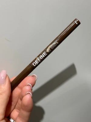 Brun ögonbrynspenna Define & Set - Endast swatchad. Snygg brun ögonbrynspenna med smal spets för exakt applicering. Pennan har en plastkropp och tydlig text i vitt. Perfekt för att forma och fylla i brynen med naturligt resultat. Pennan har en skyddande huva.