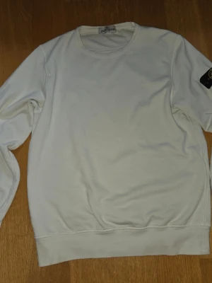 Vit sweatshirt från Stone Island - Säljer en stilren vit sweatshirt från Stone Island med klassisk rund halsringning och den ikoniska loggan på ärmen. Tröjan är i mjukt bomullsmaterial och har ribbade muddar vid ärmslut och nederkant. Perfekt för en clean och avslappnad look.