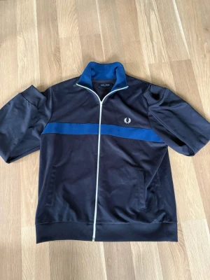 Svart track jacket från Fred Perry - Snygg svart track jacket från Fred Perry med blå rand över bröstet och vit dragkedja. Jackan har ståkrage, korta ärmar och två fickor framtill. Ikoniska Fred Perry-loggan broderad på vänster sida. Perfekt för en sportig och avslappnad stil.