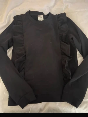 Svart sweatshirt med volangdetaljer - En svart långärmad sweatshirt med snygga volangdetaljer längs sidorna framtill. Tröjan har rund halsringning och är tillverkad i mjukt material som känns skönt mot huden. Perfekt för dig som gillar en enkel men trendig stil.