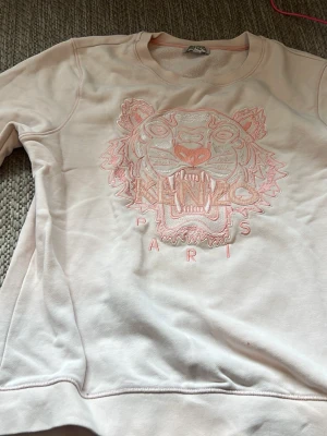 Ljusrosa Kenzo sweatshirt med tiger - Ljusrosa sweatshirt från Kenzo med ikoniskt tigerbroderi och texten 'Kenzo Paris' framtill. Tröjan har rund halsringning och långa ärmar. Perfekt för en cool och avslappnad stil.