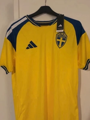 Sverige matchtröja Adidas gul/blå - Säljer en officiell gul matchtröja från Sveriges landslag med blå detaljer och tryck. Tröjan har Adidas-logga, landslagets emblem på bröstet och namn samt nummer 17 Gyökeres på ryggen. Perfekt för dig som vill stötta Sverige på match!