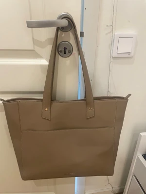 Beige handväska från Anna Field - Stilren beige handväska från Anna Field i slätt konstläder. Väskan har två långa axelband, en stor ytterficka och guldfärgade detaljer. Perfekt för dig som vill ha en enkel men snygg accessoar till din outfit.