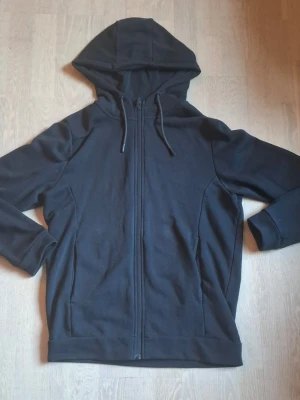 Svart zip-hoodie med huva - En stilren svart hoodie med dragkedja framtill och justerbar huva med snören. Hoodien har långa ärmar och ribbade muddar vid ärmslut och nederkant. Perfekt för en avslappnad och enkel look.