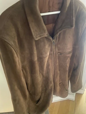 Mockajacka - stl L - XL - Säljer en brun mockajacka med klassisk krage. Jackan har dragkedja framtill, två sidofickor och en bröstficka med dragkedja. Saknas ett spänne som sitter på sidan av jackan för reglage av midjemått på vänster sida. Se bild 4.