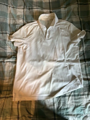 Vit kortärmad polotopp - Klassisk vit polotopp med korta ärmar och knappar vid halsen. Enkel och stilren design som passar till det mesta. Tillverkad i mjukt bomullsmaterial för en bekväm känsla.