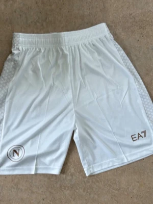 Vita EA7 shorts med logga - Snygga vita shorts från EA7 med elastisk midja och diskreta mönstrade sidopaneler. Shortsen har EA7-logga på ena benet och en rund logga på det andra. Perfekta för sommaren och sportiga outfits.