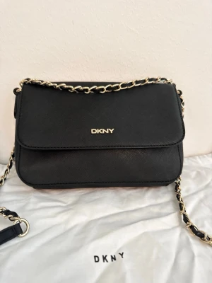 Svart axelväska från DKNY - Snygg svart axelväska från DKNY med guldfärgad kedjerem och logga framtill. Väskan har ett stilrent, rektangulärt format och är gjord i ett tåligt konstläder. Invändigt finns flera fack för bra organisering. Perfekt för dig som gillar klassisk och trendig stil.