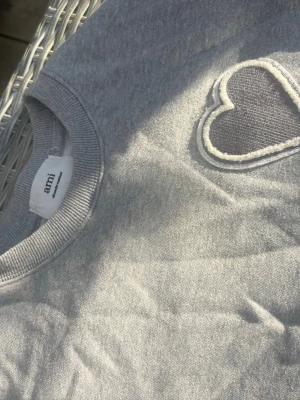 Grå sweatshirt från Ami Paris med hjärtdetalj - Säljer en stilren grå sweatshirt från Ami med deras ikoniska hjärtdetalj broderad på bröstet. Tröjan har rund halsringning och är tillverkad i mjukt bomullsmaterial. Perfekt för en avslappnad och trendig look.