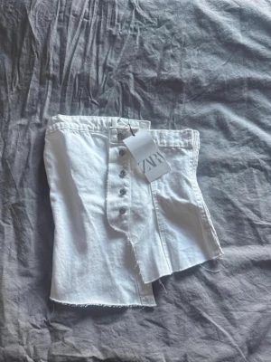 Vit denim tubtopp från Zara - Säljer en vit tubtopp från Zara i denim med rå kant nertill och knappar framtill. Helt ny med prislapp kvar. Nypris 329kr, säljer för 100kr